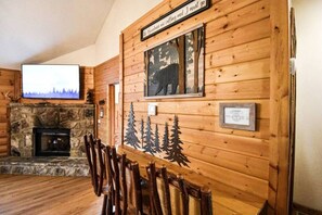 A wooden interior with a stone fireplace, wooden chairs, a television mounted on the wall, and a framed certificate.