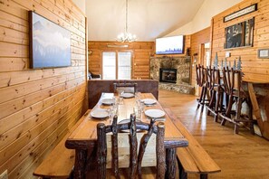 Cozy cabin dining area with a long table for four, stone fireplace, and a chandelier.