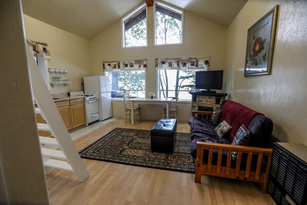 A spacious living area with a wooden floor, a black leather ottoman, a wooden coffee table, and a dark-colored sofa with cushions. There is a kitchen area with a stove, refrigerator, and a window overlooking the outdoors. - Cozy Waterfront Cabin, Central Kenai - Alaskan Sunset Cabins: 'Kalgin' (Kasilof)