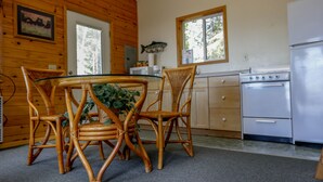 A wooden interior with a dining table surrounded by wicker chairs, a kitchen with a stove and refrigerator, and a window with a view of trees. - 'Cape Kasilof Cabin'  Waterfront/Sunset Views  (Kasilof)