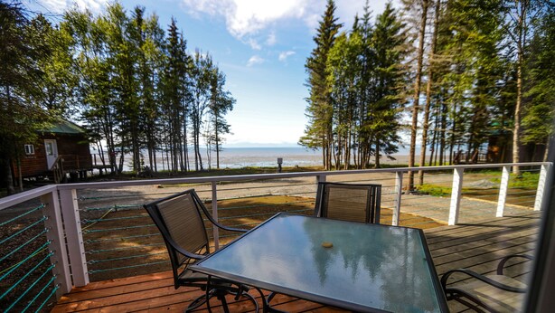 A wooden deck with a glass table and chairs, overlooking a body of water and surrounded by trees. - 'Cape Kasilof Cabin'  Waterfront/Sunset Views  (Kasilof)