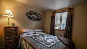A bedroom with a bed, a nightstand with a lamp, a wall decoration, and a window with curtains. - 'Cape Kasilof Cabin'  Waterfront/Sunset Views  (Kasilof)
