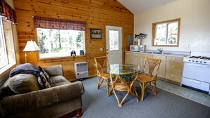 A cozy cabin-style room with a wooden interior, a glass dining table surrounded by wicker chairs, a sofa, a kitchen with a stove and microwave, and a window with a plaid valance. - 'Cape Kasilof Cabin'  Waterfront/Sunset Views  (Kasilof)