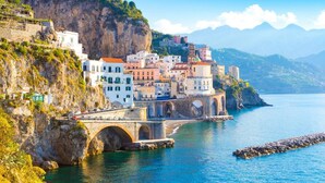 A coastal town with buildings on a cliff, a bridge, and a clear blue sea.