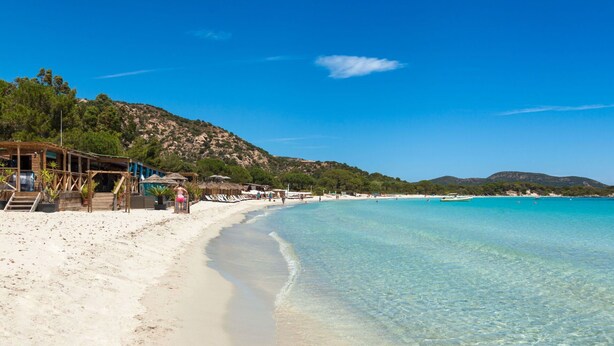 Ein Strand mit klarem türkisblauem Wasser, sandigem Ufer und Holzgebäuden mit Treppen zum Strand.