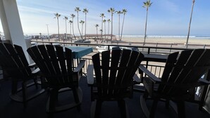 A balcony with Adirondack chairs and a table, overlooking a beach with palm trees and a parking area.