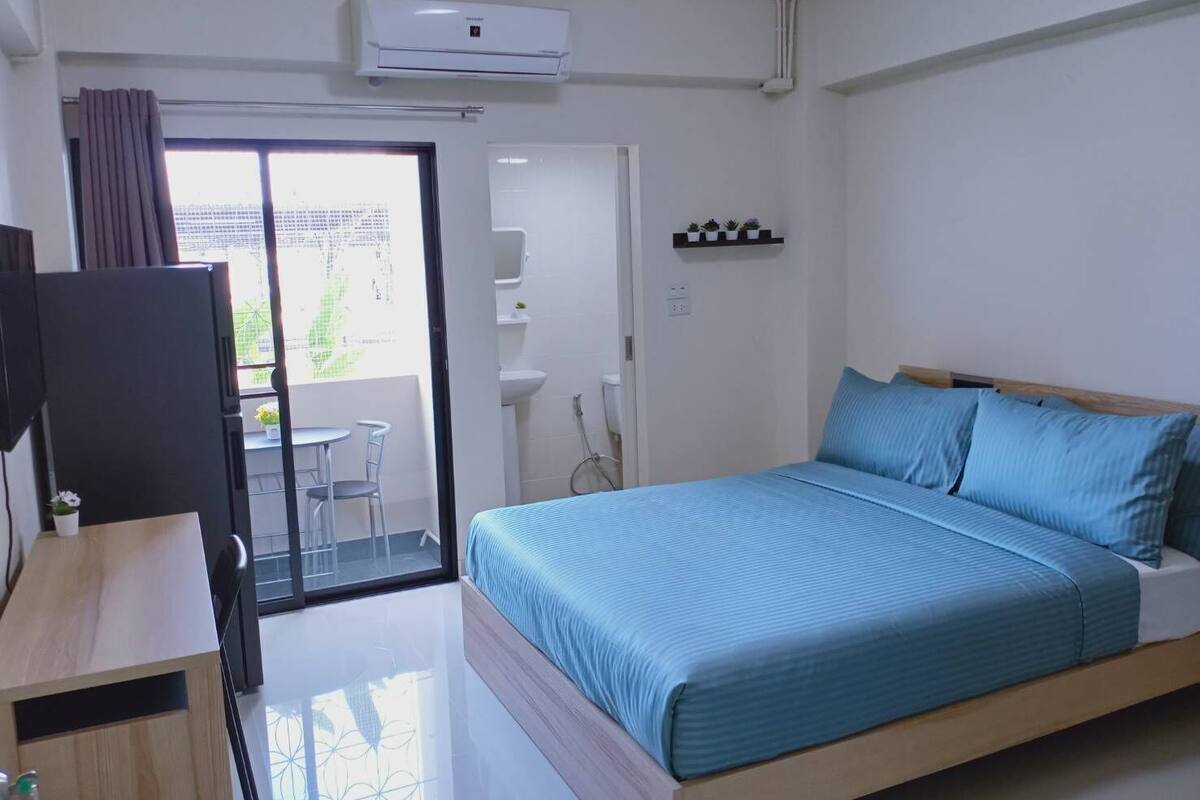Une chambre comprenant un lit, un bureau, une chaise, une télévision, un appareil de climatisation, une salle de bain avec un lavabo et une douche, et un balcon avec vue.