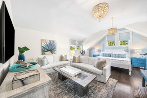 A spacious living room with a white sofa, a coffee table, and a TV. There is a ceiling fan, a chandelier, and a window with a view of greenery. - Southern Charm meets Modern Luxury | Electric Cart, Private Pool, Bikes, & Beach Club Access (Santa Rosa Beach)