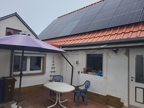 A house with a solar panel roof, a patio with a table and chairs, and a small window with a plant.