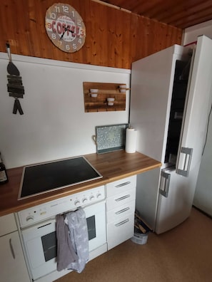 A kitchen with a wooden wall, a clock, a stove, a refrigerator, and a wooden shelf with cups.