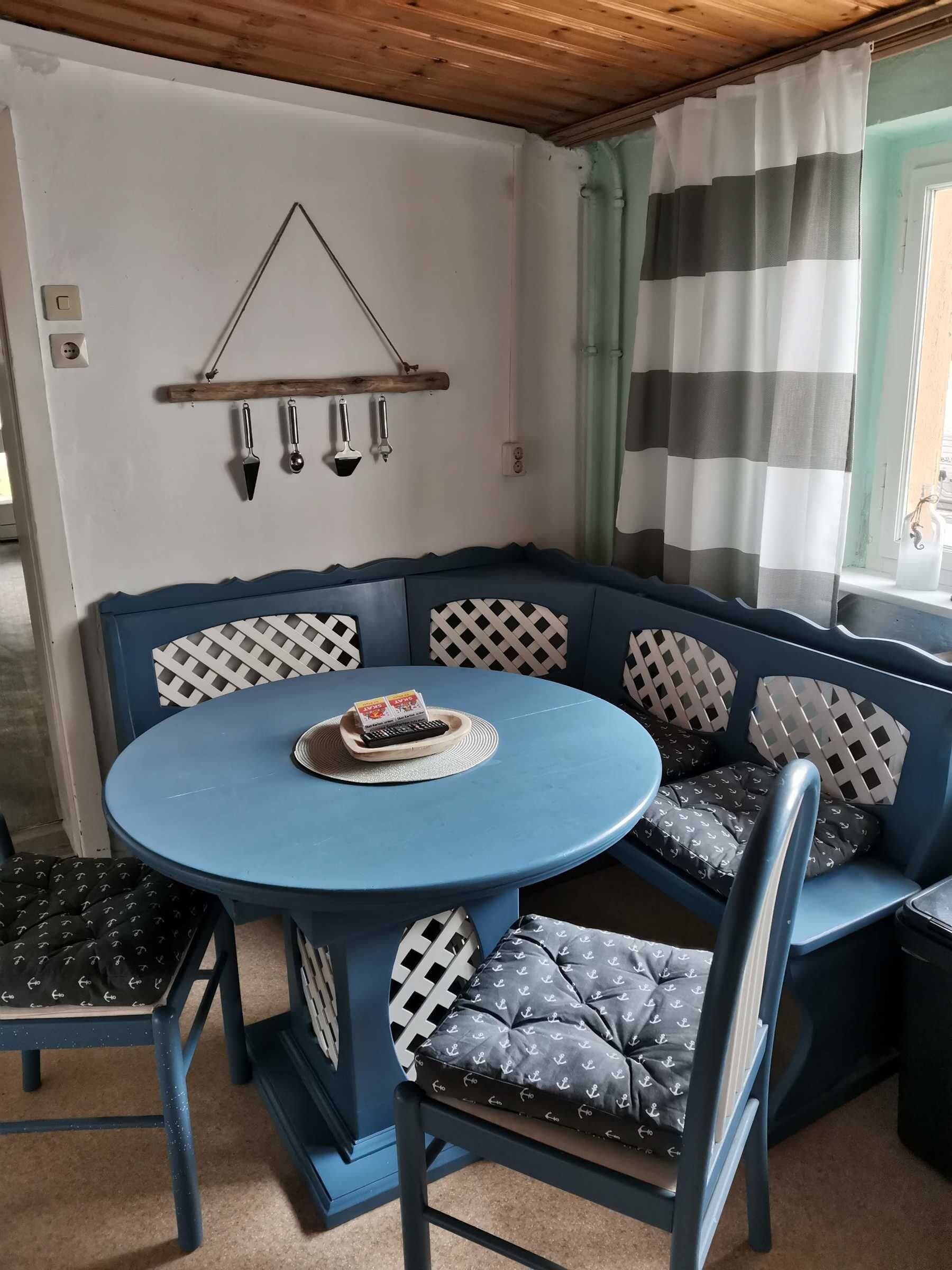 Un canapé d’angle bleu avec des coussins à motifs, une petite table ronde avec une télécommande, et une étagère en bois contenant des ustensiles de cuisine.