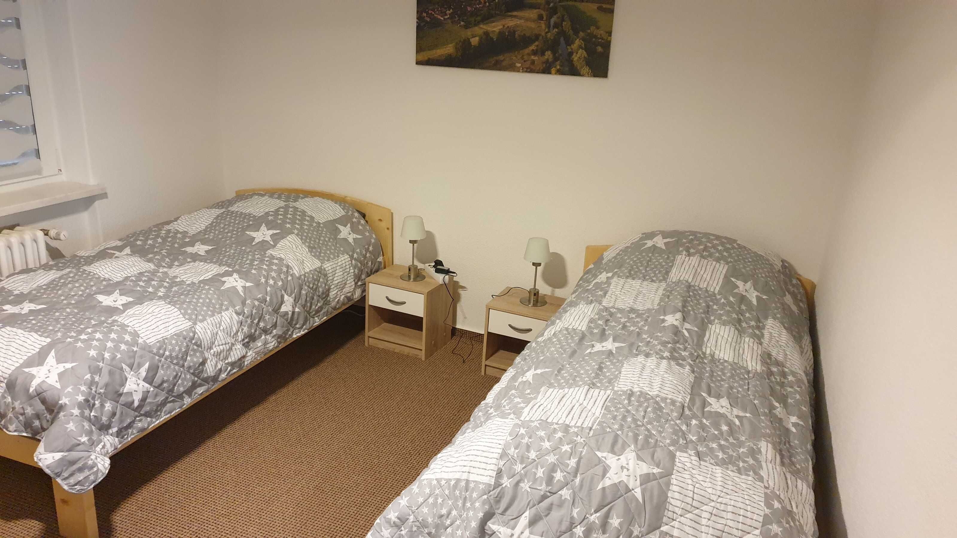 A bedroom with two single beds, each with a star-patterned bedspread, wooden headboards, bedside tables with lamps, and a radiator on the wall.