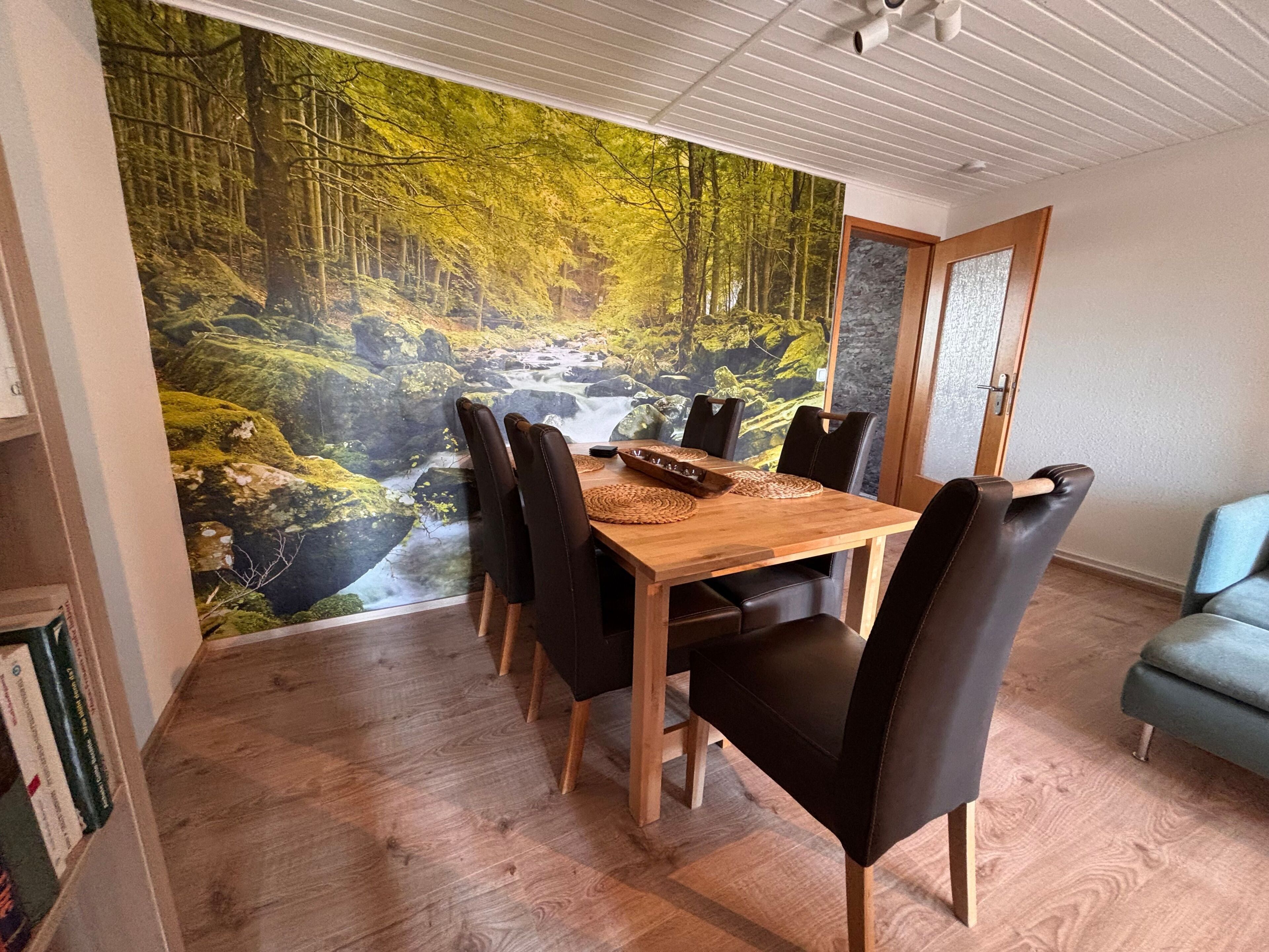 Ein Essbereich mit Holztisch, schwarzen Stühlen, einer Waldmalerei und einem blauen Sofa.