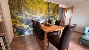 A dining area with a wooden table, black chairs, a forest mural, and a blue sofa. - Apartment Hohensee ground floor - Ferienwohnung Hohensee (Clausthal-Zellerfeld)