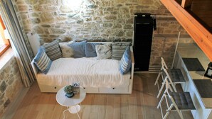 Ein gemütliches Zimmer mit Steinwand, einem Bett mit blau-weißer Bettwäsche, einem kleinen runden Tisch mit einer Pflanze und einer Treppe mit grau gepolsterten Stufen.