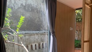 A room with a large window, wooden flooring, and a view of a stone wall and greenery outside. - Hip's House Homestay - Hostel (Phu Quoc)