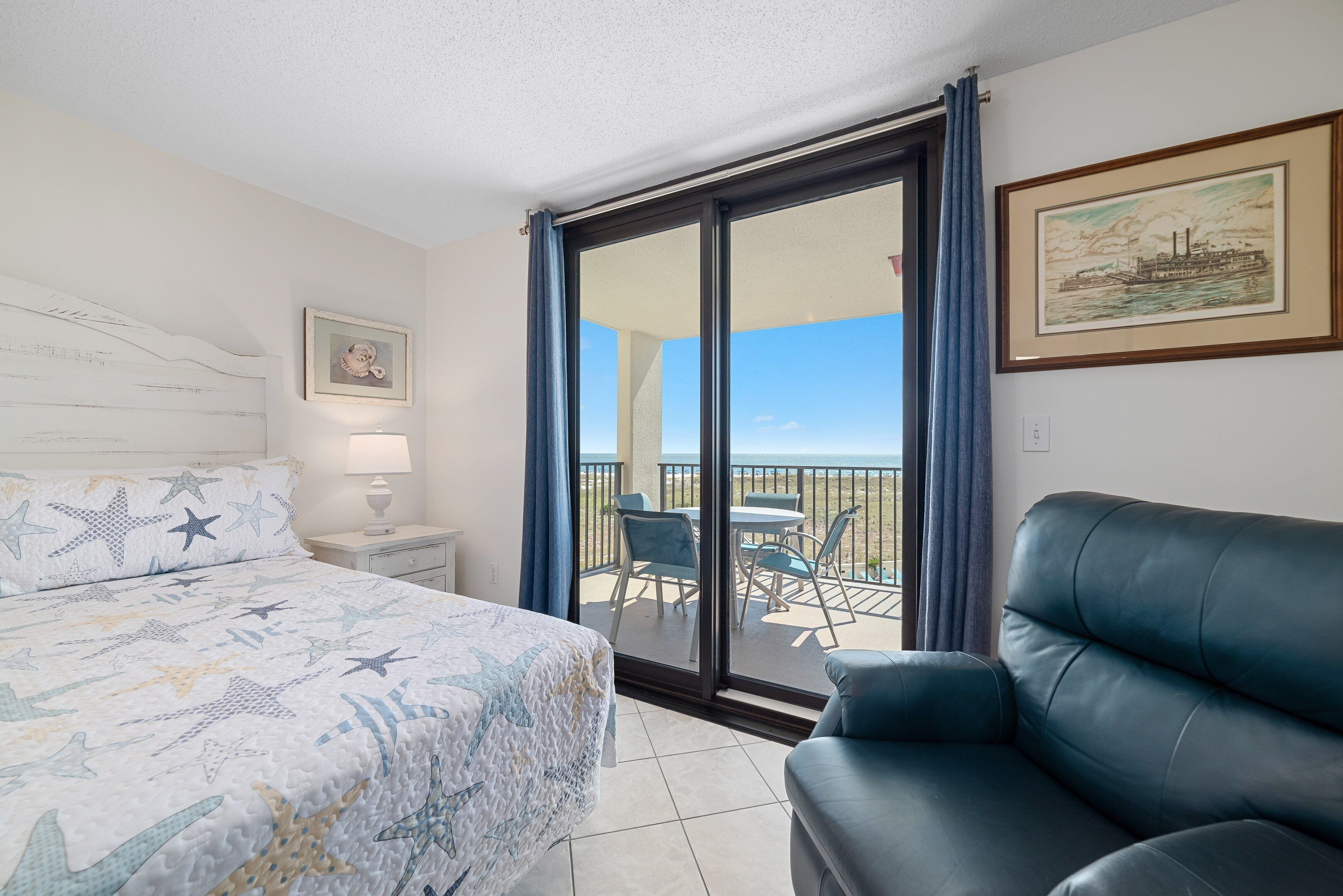 A hotel room with a bed, a blue leather sofa, a balcony with a table and chairs, and a painting on the wall.