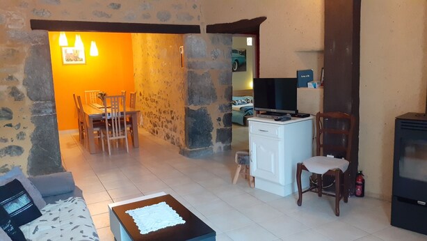Un salon chaleureux avec un mur en pierre, un coin repas avec des chaises, une petite table recouverte d’une nappe blanche, un téléviseur et une cheminée.