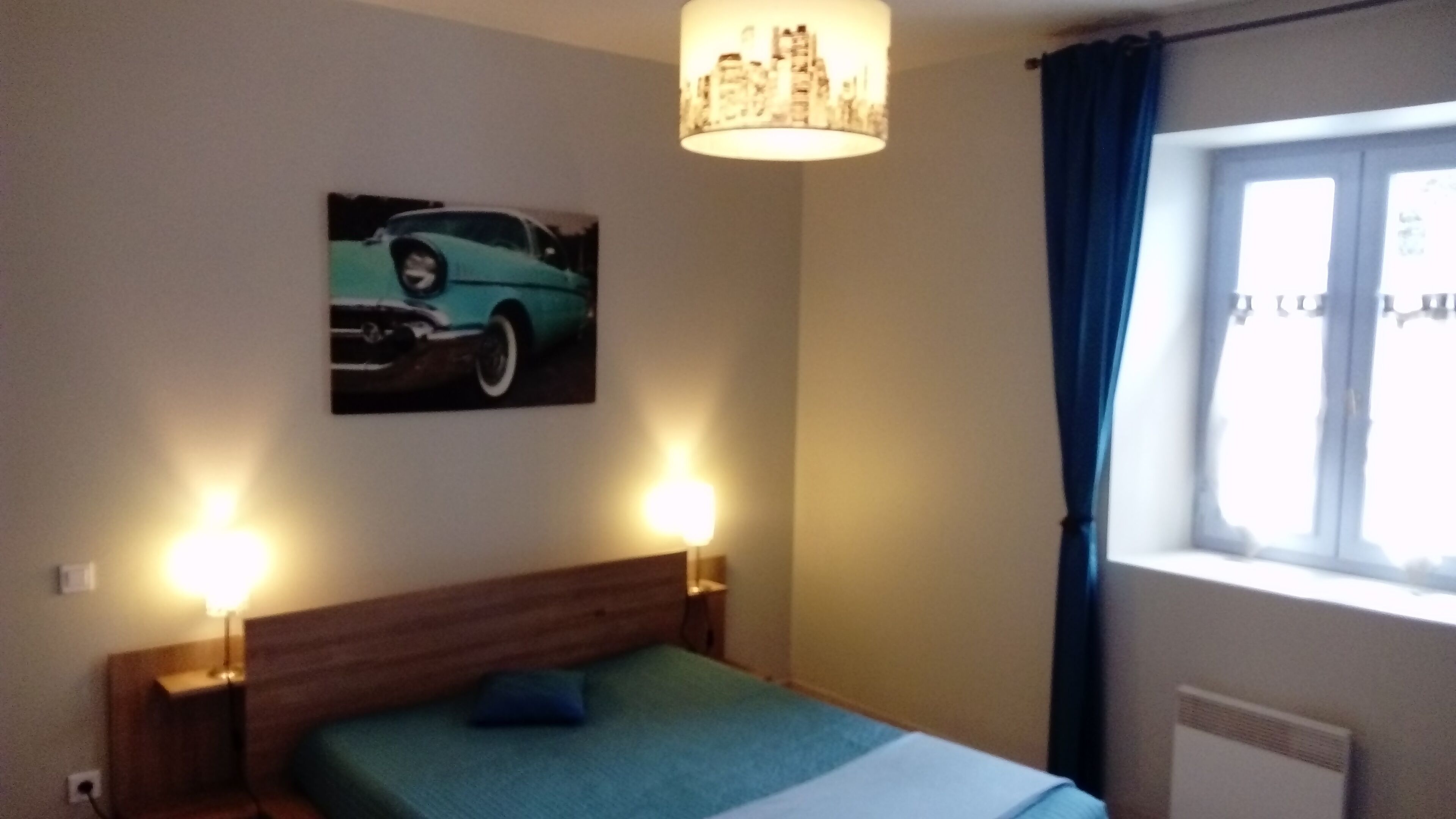 Une chambre à coucher avec un lit, un tableau représentant un paysage urbain, deux lampes de chevet, une fenêtre avec des rideaux et un luminaire au plafond.