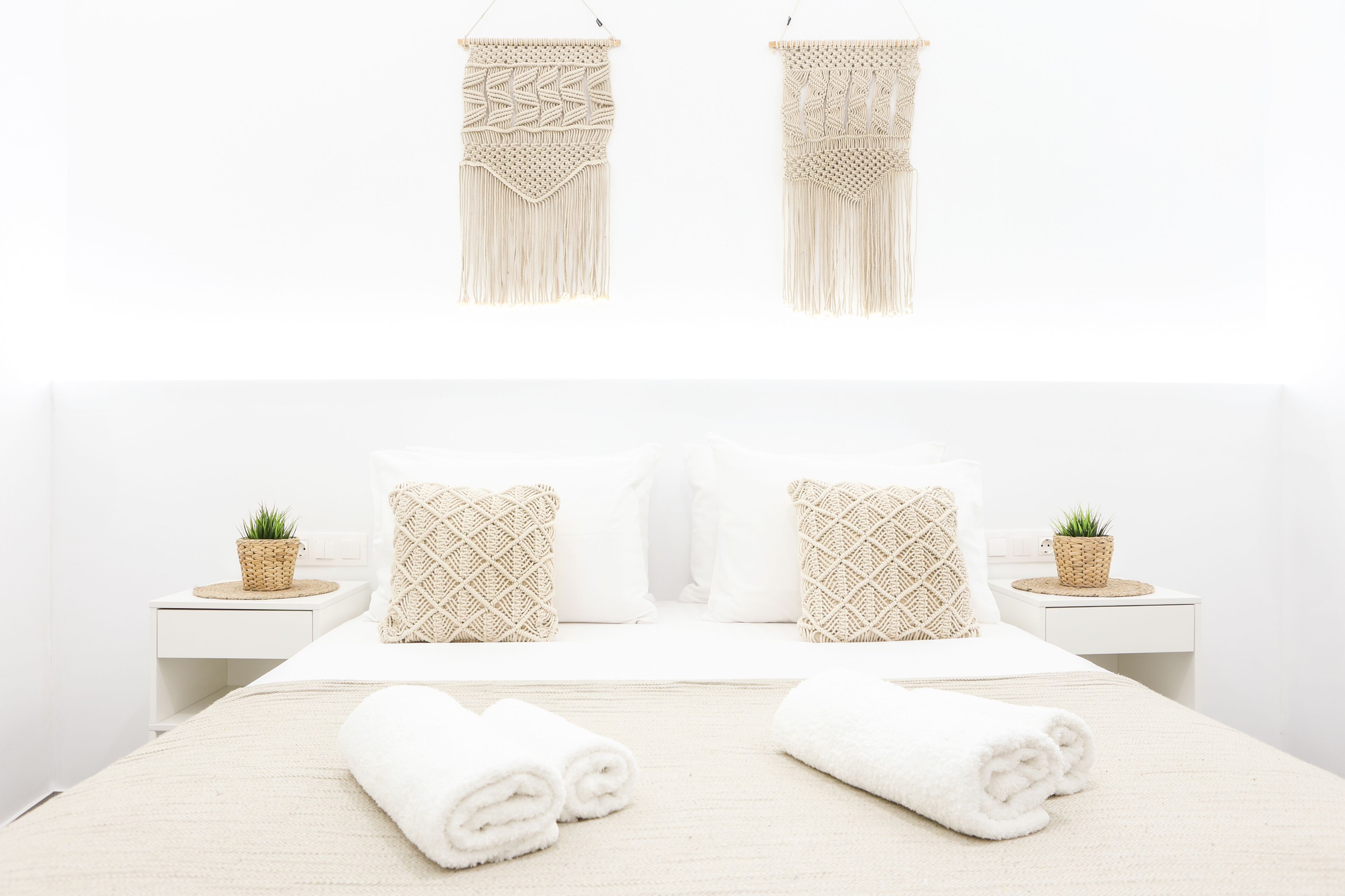Una cama bien hecha con dos almohadas, dos mesitas de noche con pequeñas plantas, y dos tapices de macramé en la pared.
