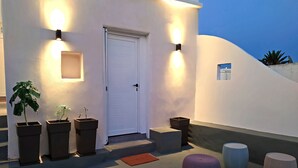 Fachada de edificio con una puerta blanca, dos luces fijadas a la pared y plantas en macetas.