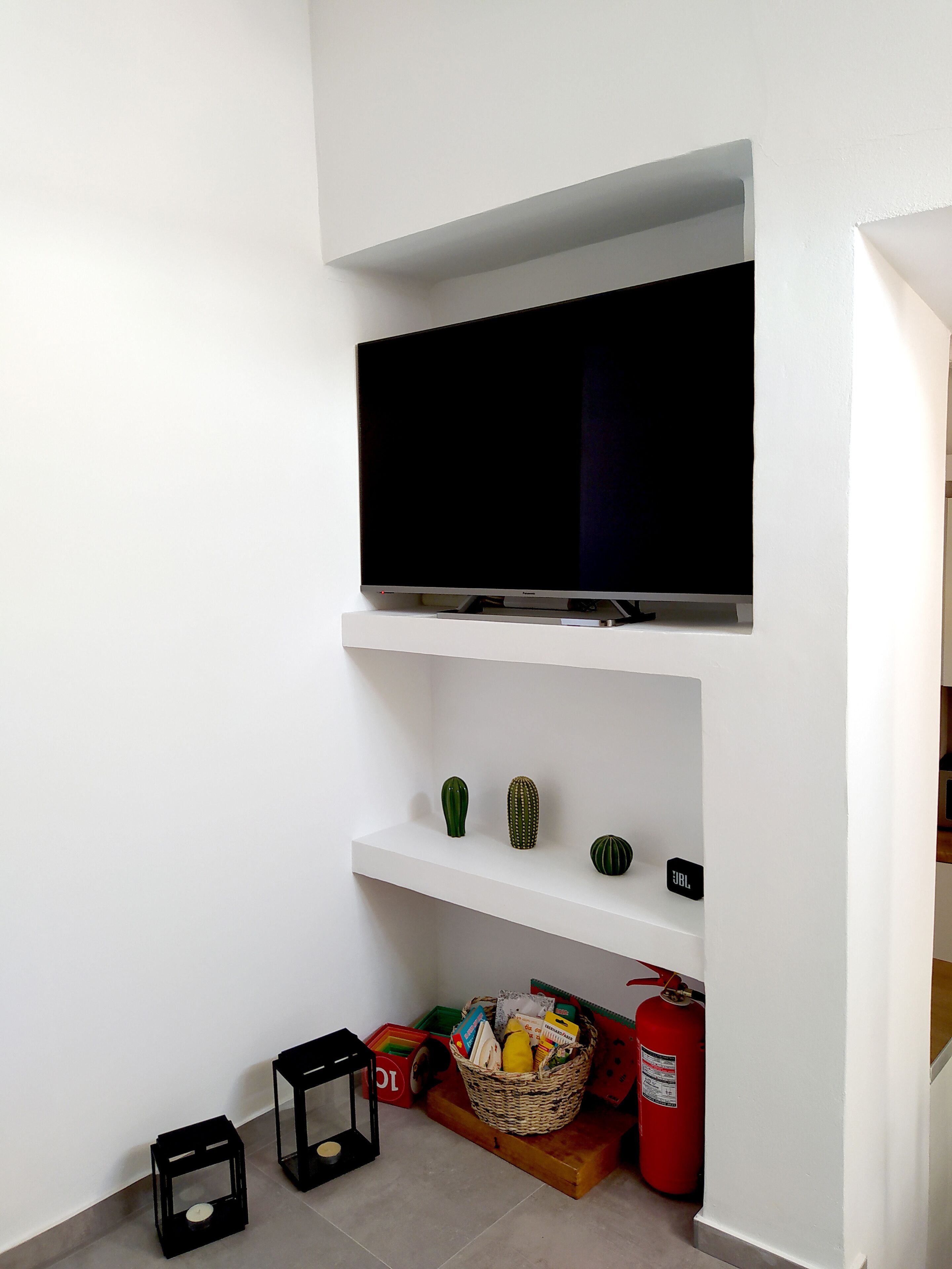 A room with a mounted flat-screen TV, a shelf with decorative items, a basket with groceries, a fire extinguisher, and a small table with a candle.