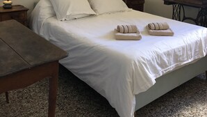 Ein Einzelbett mit weißen Leinentüchern und zwei aufgerollten Handtüchern, ein Nachttisch aus Holz mit einer Lampe sowie ein kleiner Tisch ebenfalls mit einer Lampe.