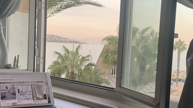 Ein Zimmer mit großem Fenster mit Blick auf einen Strand, ein Laptop auf einem Schreibtisch und ein kleiner, runder Hocker.