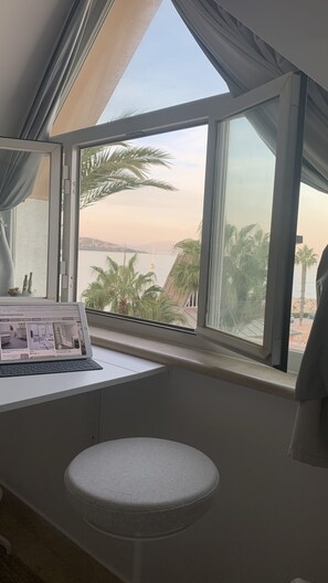 Quarto com uma janela grande com vista para uma praia, um laptop sobre uma mesa e um banquinho redondo.