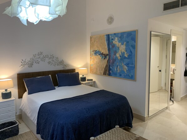 A bedroom with a large bed, a nightstand, a wall-mounted lamp, a decorative wall piece, and a door leading to another room.