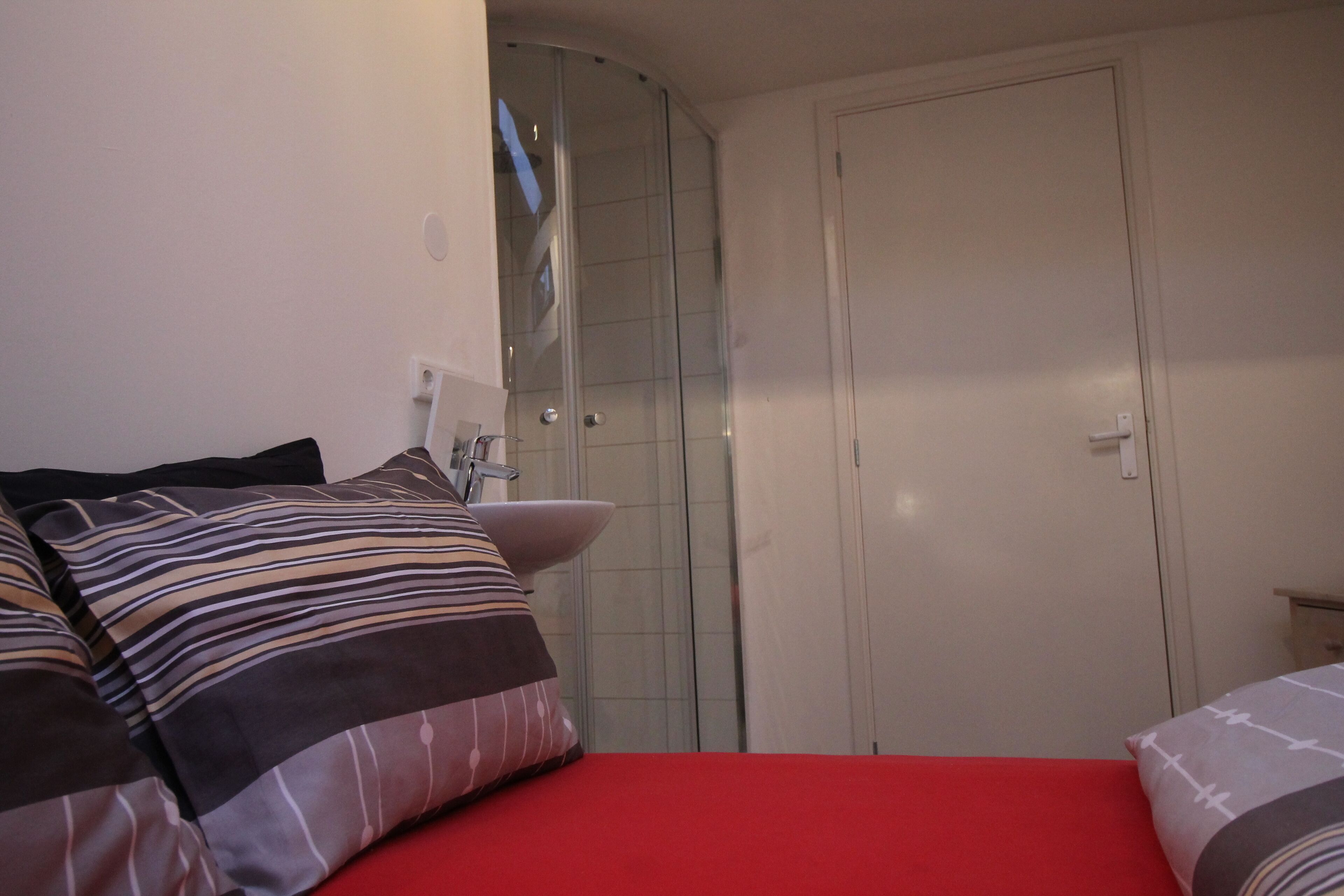 A bedroom with a red bedspread, a white door, and a shower area.