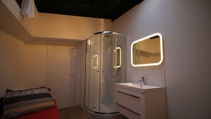 Een moderne badkamer met een douchecabine van glas, een wastafel met spiegel en een rode bed met gestreepte kussens.