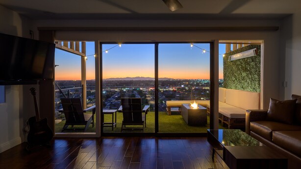 A modern living room with a large sliding glass door leading to an outdoor patio. The room features a wooden floor, a leather sofa, a flat-screen TV, and a guitar. The patio has a fire pit and outdoor seating.