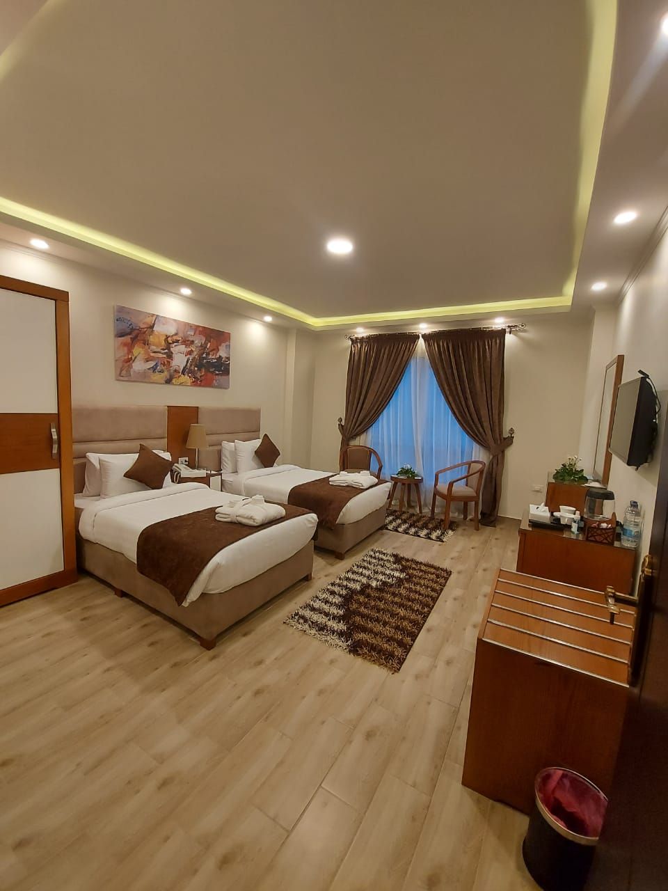 A hotel room with two beds, a desk, a chair, a TV, and a window with curtains.