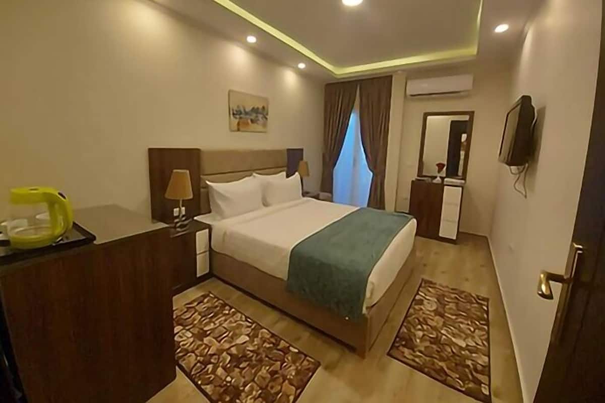 A hotel room with a bed, bedside tables, a desk with a lamp, a TV, and an air conditioning unit.