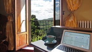 Ein Laptop auf einem Tisch, dazu eine Tasse und ein Untertasse, ein Fenster mit Vorhängen und Blick ins Grüne.