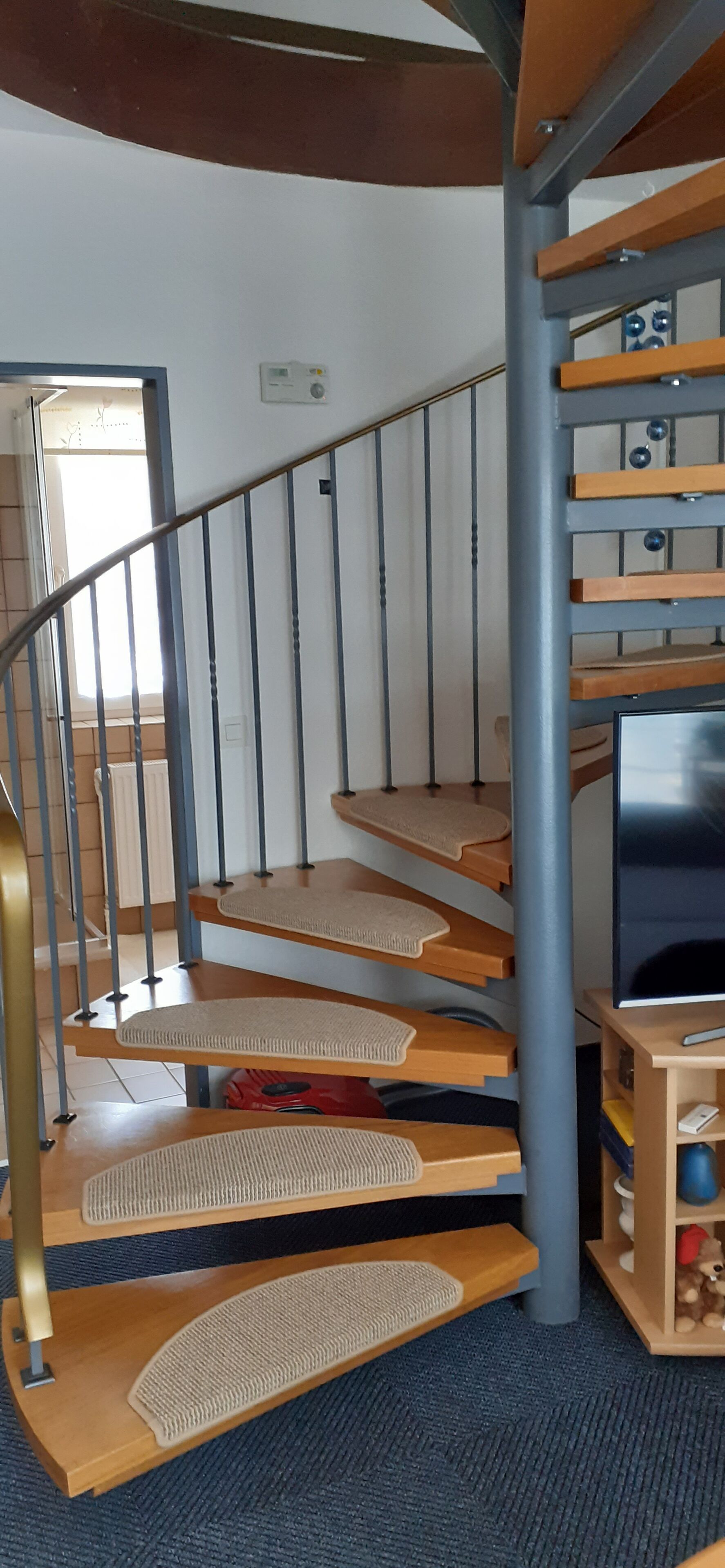 Un escalier en colimaçon avec des marches en bois et des rampes en métal, menant à un palier où se trouvent un téléviseur et une petite étagère avec quelques objets.