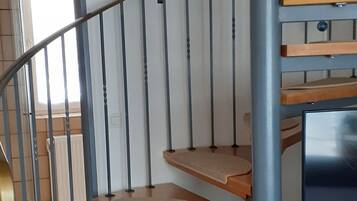 Un escalier en colimaçon avec des marches en bois et des rampes en métal, menant à un palier où se trouvent un téléviseur et une petite étagère avec quelques objets.