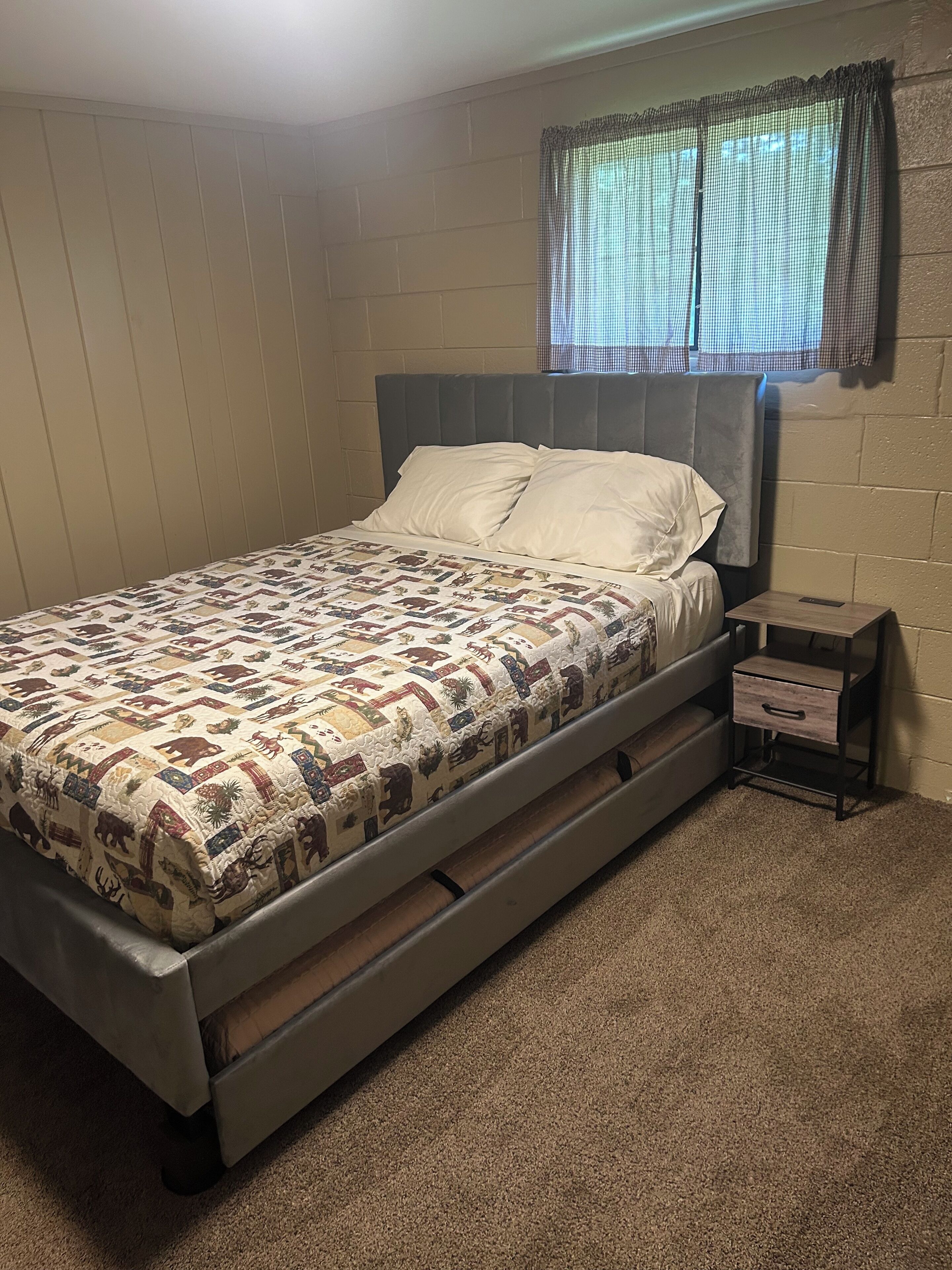 A single bed with a quilt, a nightstand with a drawer, and a window with a checkered curtain.