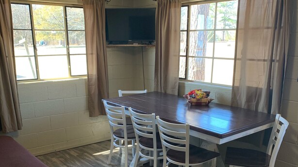 Una habitación con una mesa de comedor de madera, seis sillas blancas, un televisor montado en la pared y una cama con estructura de madera.