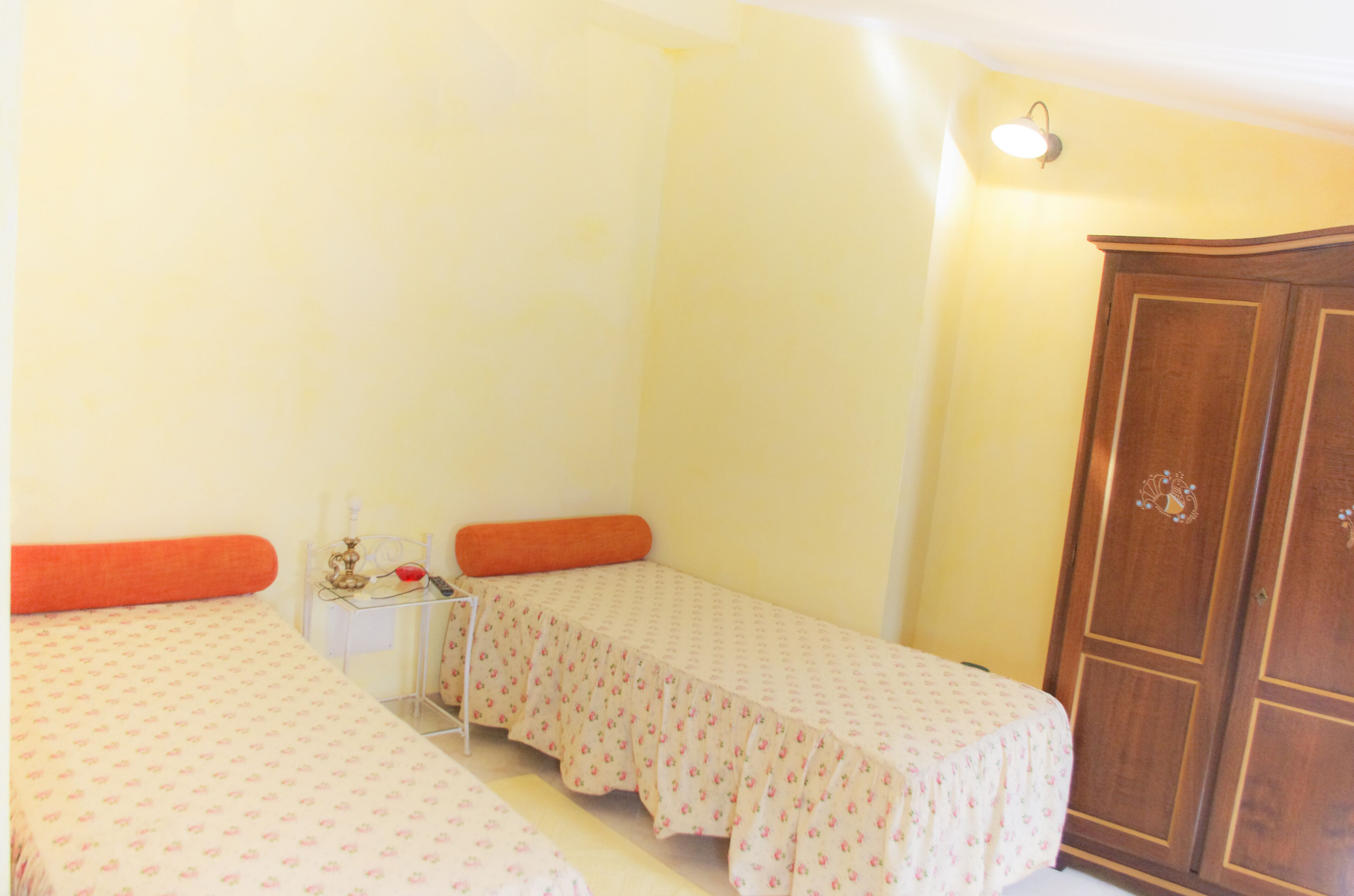 A small, single-room accommodation with two single beds, a wooden wardrobe, and a bedside table with a lamp.