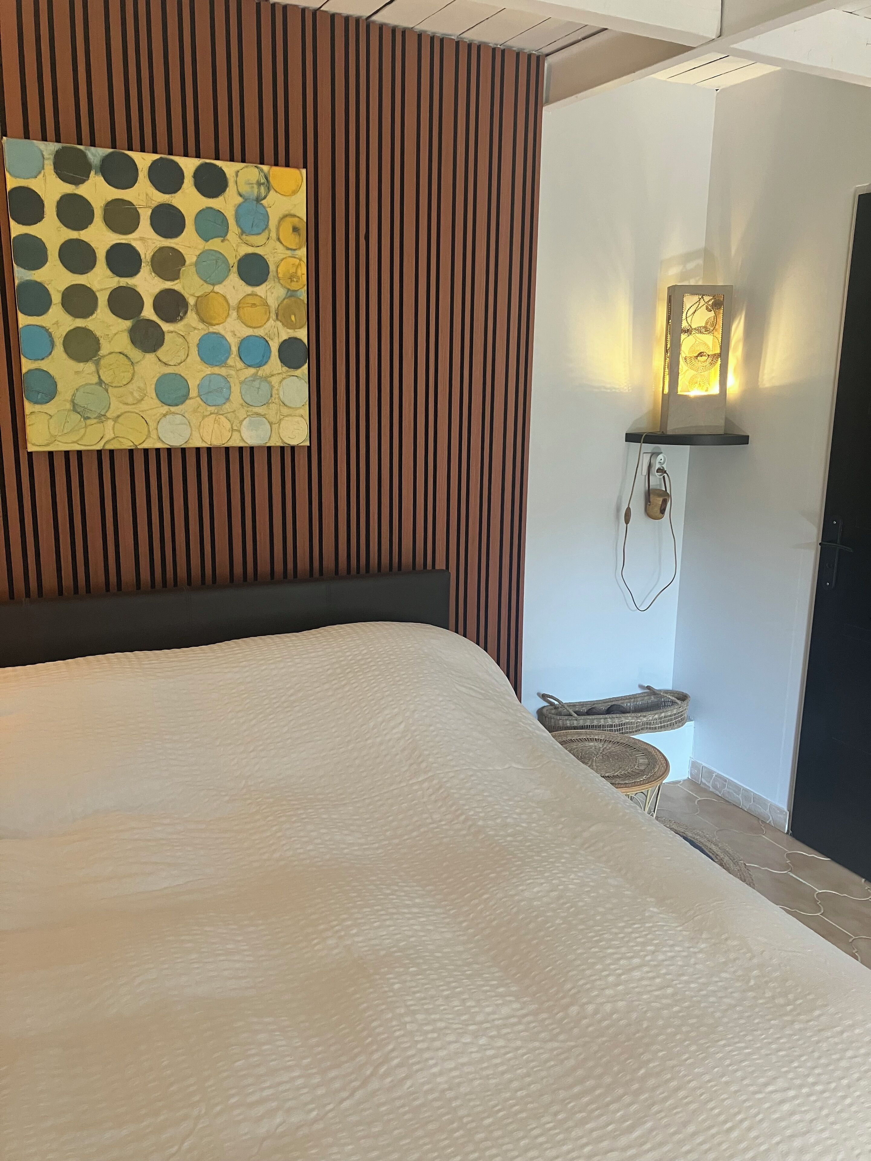 Una cama bien hecha con una cubierta blanca texturizada, una obra de arte colgada en la pared con un estampado de lunares y una lámpara de pared con pantalla decorativa.