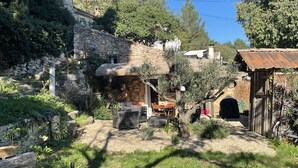 Edifici de pedra rústic amb taula i cadires de fusta a l'exterior, envoltat de vegetació i una bicicleta.
