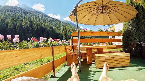 Une terrasse en bois, dotée de bancs et d’un parasol, qui offre une vue sur un jardin fleuri et sur une chaîne de montagnes en arrière-plan.