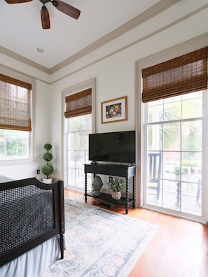 A room with a large window, a ceiling fan, a television on a stand, and a small potted plant.