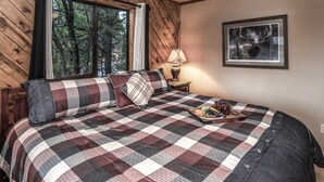Una camera da letto con un letto grande, biancheria a quadretti, un comodino con una lampada, un quadro appeso al muro e una finestra vista alberi.