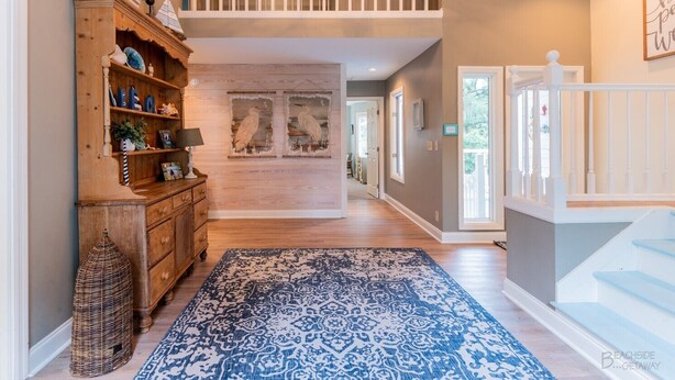 Un hall d’entrée spacieux avec un plancher en bois, un tapis à motifs bleus, une bibliothèque en bois et un escalier avec des rampes blanches.