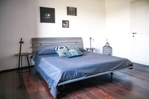 Un lit bien fait, avec un matelas gris et des coussins à motifs, une petite table ronde surmontée d’une lampe, et une cage à oiseaux décorative posée sur le sol.