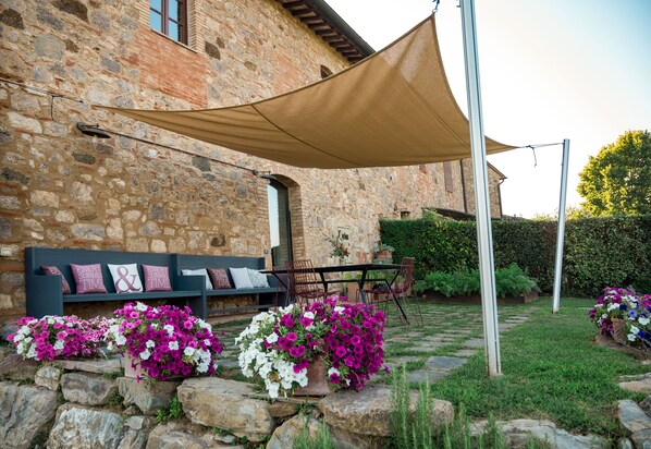 Un bâtiment en pierre avec un banc en pierre et une table entourée de chaises. On y trouve des pots de fleurs contenant des fleurs roses et blanches.