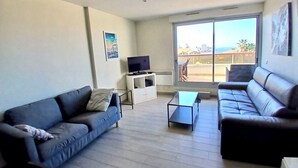 Een woonkamer met een bank, een salontafel en een tv op een standaard. Er is een balkon met uitzicht op de stad.